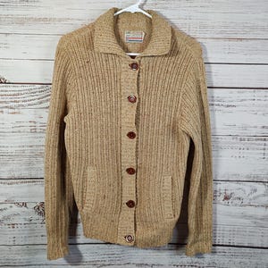 May include: A tan cardigan sweater with a button-up front and a ribbed texture. The sweater has a collared neckline and two pockets.