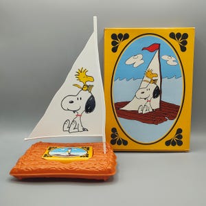 May include: A Snoopy-themed toy sailboat with a white sail featuring Snoopy and Woodstock. The boat sits on an orange base. A matching box with a similar illustration is next to the boat. The box has a yellow border.
