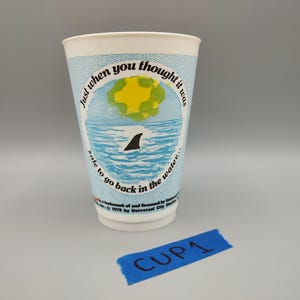 May include: A white paper cup with a light blue background and a graphic of a shark fin in the ocean. The cup has the text "Just when you thought it was safe to go back in the water." The cup is labeled "CUP1".