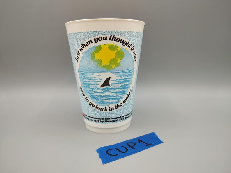 May include: A white paper cup with a light blue background and a graphic of a shark fin in the ocean. The cup has the text "Just when you thought it was safe to go back in the water." The cup is labeled "CUP1".