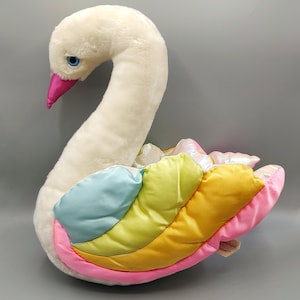 May include: A plush swan toy with soft white fur, a bright pink satin beak, and blue eyes. The wings are constructed from padded, multi-colored satin fabric panels in shades of light blue, lime green, yellow, and pink, featuring a subtle iridescent sheen.