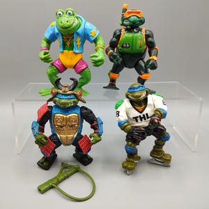 May include: Four Teenage Mutant Ninja Turtles action figures. One is a frog with a blue jacket, another is wearing a diving mask, a third is in samurai armor, and the last is on ice skates. A green slingshot is in the foreground.
