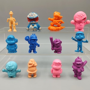 K&ouml;nnte beinhalten: Eine Sammlung bunter, kleiner Figuren in verschiedenen Posen. Die Figuren sind in Blau-, Rosa-, Orange- und Lilat&ouml;nen gehalten. Einige Figuren sind Zeichentrickfiguren, andere haben die Form von Menschen. Die Figuren werden auf einem transparenten, mehrstufigen Regal ausgestellt.