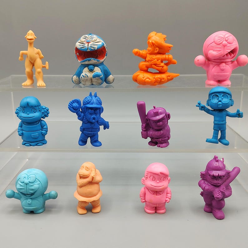 K&ouml;nnte beinhalten: Eine Sammlung bunter, kleiner Figuren in verschiedenen Posen. Die Figuren sind in Blau-, Rosa-, Orange- und Lilat&ouml;nen gehalten. Einige Figuren sind Zeichentrickfiguren, andere haben die Form von Menschen. Die Figuren werden auf einem transparenten, mehrstufigen Regal ausgestellt.