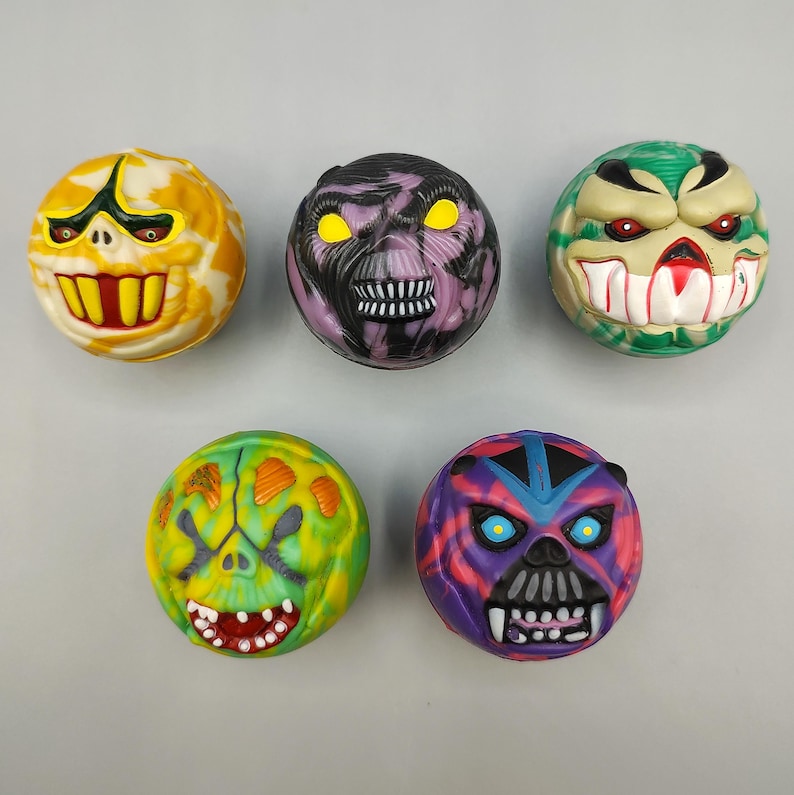 May include: Five colorful, round toys with monster faces. The toys feature various colors like yellow, green, purple, and red, with detailed facial features including teeth, eyes, and unique designs. These toys are likely collectible or novelty items.
