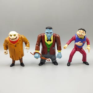 May include: Three colorful action figures of characters from a movie. The figures include a character in a tan coat, a blue-skinned character in a brown jacket, and a character in a red suit. The figures are standing on a gray surface.