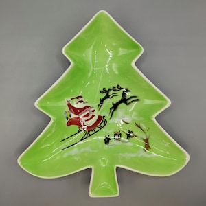May include: A green, Christmas tree-shaped serving dish with a white rim. The dish features a painted scene of Santa Claus in his sleigh being pulled by reindeer. The design includes gifts and a festive holiday theme.