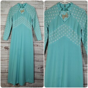 May include: A long, turquoise blue dress with white floral embroidery on the sleeves and neckline. The dress has a high neckline and a bow detail at the neckline.