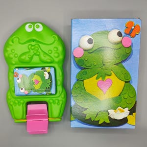 May include: A green plastic frog-shaped toy with a pink lever and a small window displaying a frog puzzle. A matching box features a frog illustration with a heart, butterfly, and flower.