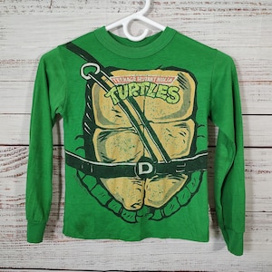May include: Green long-sleeve shirt featuring a turtle shell design with the words "Teenage Mutant Ninja Turtles" in yellow and green. The shirt has a crew neck and is hanging on a white hanger.