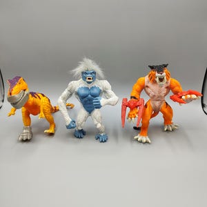 May include: Three colorful action figures: a yellow and orange dinosaur, a white and blue yeti, and an orange and tan tiger-like creature. Each figure has unique details and poses, suggesting a fantasy or adventure theme.