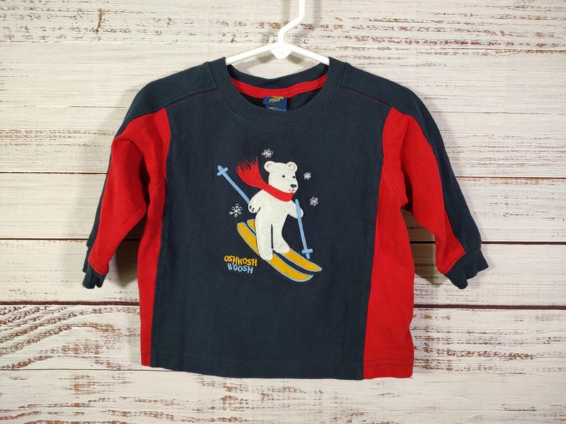May include: Navy blue and red long-sleeved top with a white bear skiing. The bear wears a red scarf and holds blue ski poles. The top has the text "OSHKOSH B'GOSH" and is hanging on a white hanger.