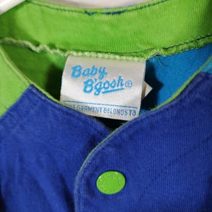 May include: Close-up of a blue, green, and white baby's shirt. The shirt has a green collar and a blue body with a green button. The tag reads "Baby B'gosh".