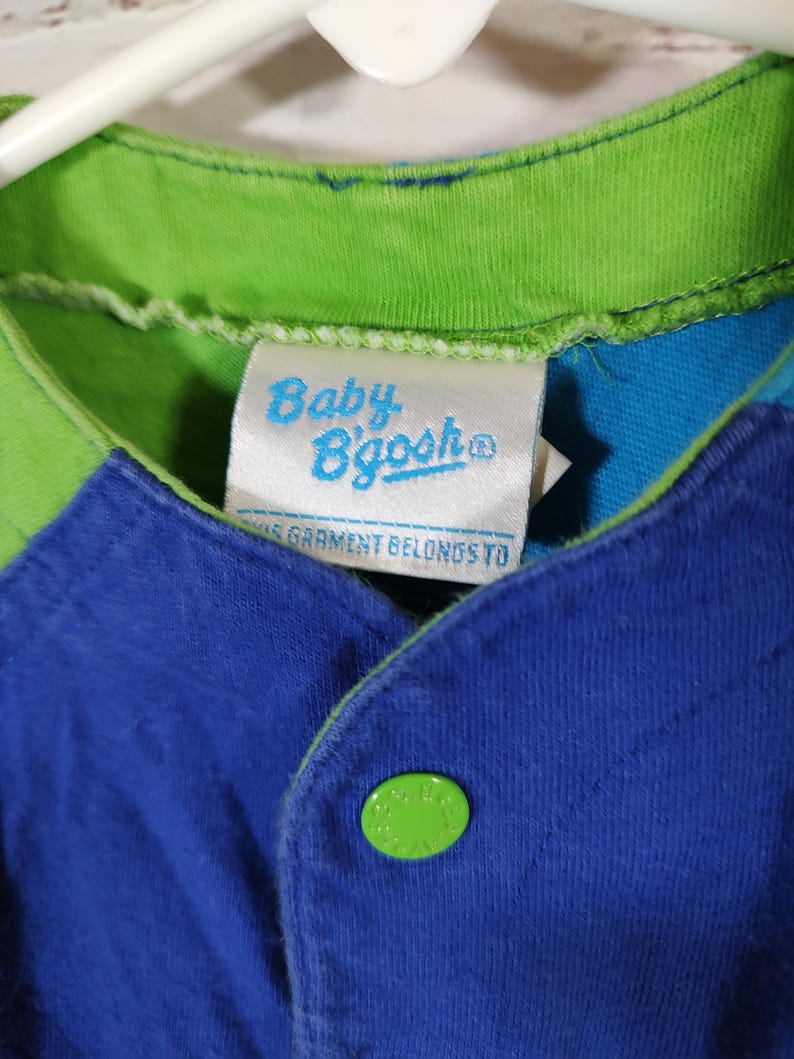 May include: Close-up of a blue, green, and white baby's shirt. The shirt has a green collar and a blue body with a green button. The tag reads "Baby B'gosh".