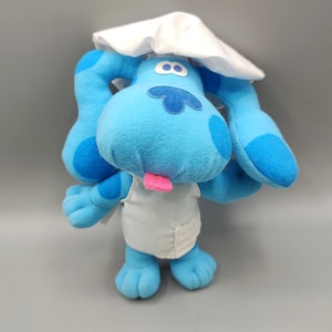May include: A plush blue dog toy with large ears and a pink tongue sticking out. The dog is wearing a white chef's hat and apron. The toy has dark blue spots and a light blue nose.