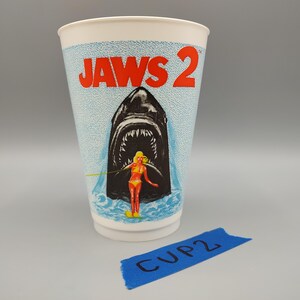 May include: A white paper cup with the words "JAWS 2" in red. The cup features an illustration of a shark's open mouth with a woman in a yellow bikini. The cup has a blue and white speckled background.