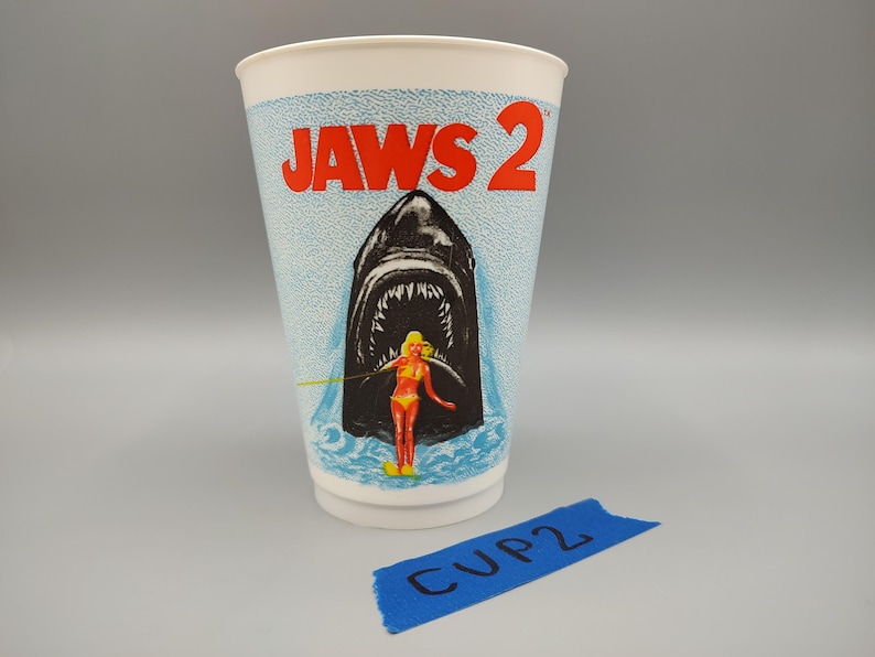 May include: A white paper cup with the words "JAWS 2" in red. The cup features an illustration of a shark's open mouth with a woman in a yellow bikini. The cup has a blue and white speckled background.
