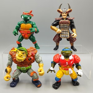 May include: Four Teenage Mutant Ninja Turtles action figures. One is green with an orange mask, another is a samurai, a third is a mutant, and the fourth is wearing a football uniform. The figures are on a clear display shelf.