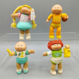 May include: Four vintage Cabbage Patch Kids dolls. One doll has blonde pigtails and a pink and blue dress, holding a bottle. Another doll is wearing a yellow football uniform and holding a football. The other two dolls are wearing baby clothes.