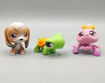 Puede incluir: Tres figuras de animales coleccionables y coloridas. Un perro marrón y blanco con ojos grandes, una tortuga verde con caparazón naranja y una criatura rosa con un lazo amarillo.