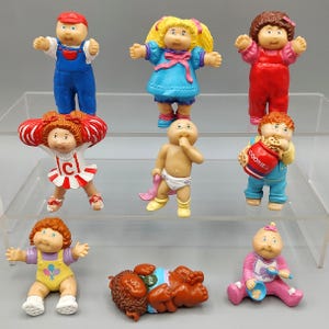 May include: A collection of vintage Cabbage Patch Kids figurines. The figurines are in various poses and outfits, including overalls, dresses, and a diaper. The colors are bright and varied, with a focus on primary colors and pastels. Some figurines are holding accessories.