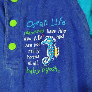 May include: Blue baby's overall with green buttons. The text "Ocean Life" is embroidered on the front, along with a seahorse illustration and the phrase "seahorses have fins and gills and are not really horses at all." The brand name "baby b'gosh" is also visible.