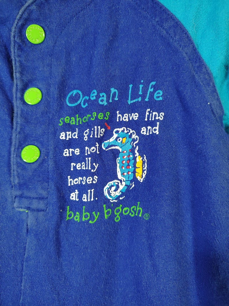 May include: Blue baby's overall with green buttons. The text "Ocean Life" is embroidered on the front, along with a seahorse illustration and the phrase "seahorses have fins and gills and are not really horses at all." The brand name "baby b'gosh" is also visible.