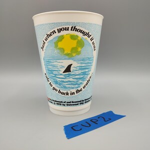 May include: A white paper cup with a light blue band featuring a shark fin in the ocean, a yellow sun, and the text "Just when you thought it was safe to go back in the water." A blue label reads "CUP 2."