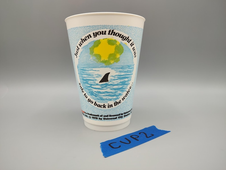 May include: A white paper cup with a light blue band featuring a shark fin in the ocean, a yellow sun, and the text "Just when you thought it was safe to go back in the water." A blue label reads "CUP 2."