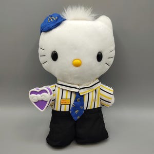 May include: A white plush Hello Kitty doll wearing a blue McDonald's uniform hat and a striped shirt. The doll holds a purple heart-shaped box and has a blue tie with the McDonald's logo. The text "Dear Daniel" is on the shirt.