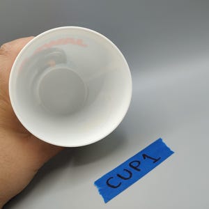 May include: A white cup with a rounded base, viewed from above. The interior is white, and the rim has a faint red design. A piece of blue tape with "CUP 1" written on it is in the lower right corner.