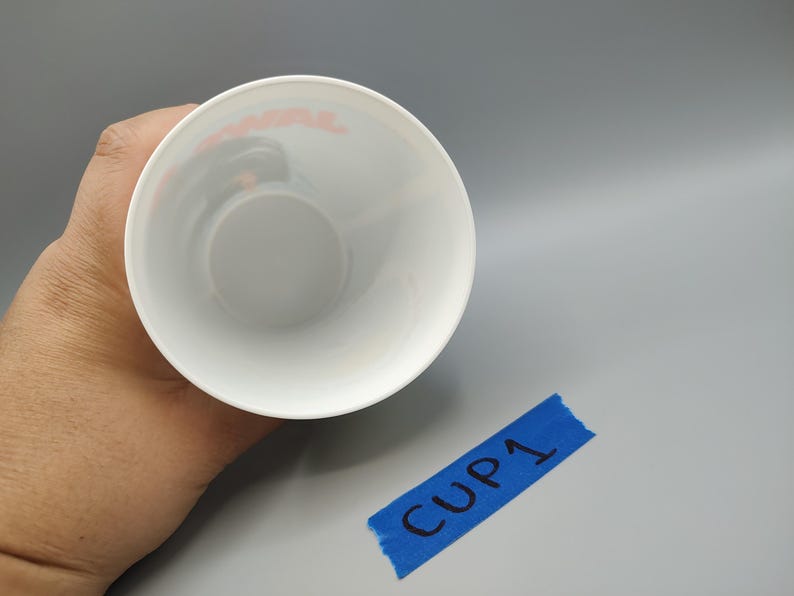 May include: A white cup with a rounded base, viewed from above. The interior is white, and the rim has a faint red design. A piece of blue tape with "CUP 1" written on it is in the lower right corner.
