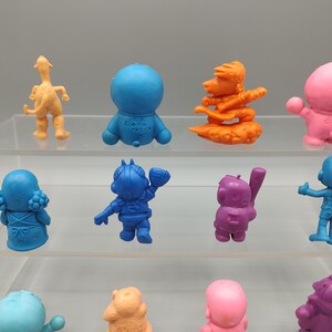 K&ouml;nnte beinhalten: Eine Sammlung farbenfroher, kleiner Figuren in verschiedenen Posen. Die Figuren sind in Blau-, Orange-, Rosa- und Lilat&ouml;nen gehalten. Einige stehen, andere befinden sich in Aktionsposen. Die Figuren werden auf einem transparenten, mehrstufigen Regal ausgestellt.