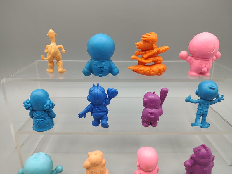 K&ouml;nnte beinhalten: Eine Sammlung farbenfroher, kleiner Figuren in verschiedenen Posen. Die Figuren sind in Blau-, Orange-, Rosa- und Lilat&ouml;nen gehalten. Einige stehen, andere befinden sich in Aktionsposen. Die Figuren werden auf einem transparenten, mehrstufigen Regal ausgestellt.