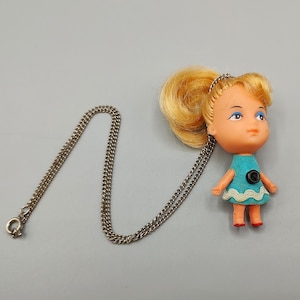 May include: A necklace featuring a small doll pendant with blonde hair, blue eyeshadow, and a turquoise dress with a white wavy trim and a black button detail. The doll is attached to a silver-toned metal chain with a circular clasp closure.
