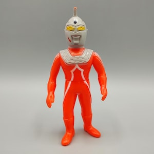 May include: A vintage action figure of a superhero in a bright orange suit with white accents. The figure has a gray helmet with yellow and white details. It stands upright with arms at its sides, showcasing a classic design.
