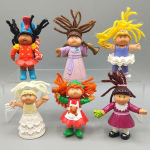May include: A collection of six Cabbage Patch Kids figurines. Each doll has yarn hair and is dressed in a unique outfit, including a soldier, angel, and ballerina. The dolls are made of plastic and have painted features.