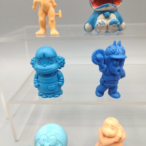 K&ouml;nnte beinhalten: Eine Sammlung bunter, kleiner Plastikfiguren. Die Figuren haben verschiedene Formen, darunter Zeichentrickfiguren und menschliche Figuren. Die Farben sind Blau, Orange und Beige. Die Figuren werden auf einem transparenten, mehrstufigen Regal ausgestellt.