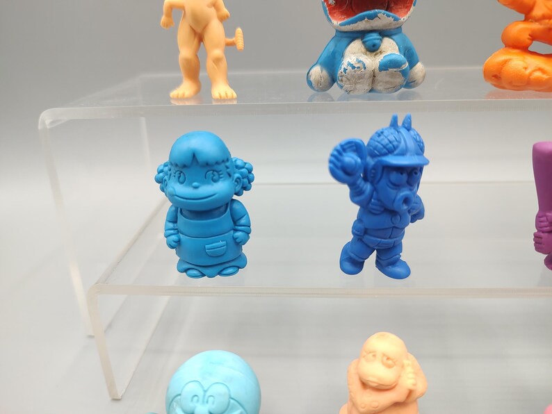 K&ouml;nnte beinhalten: Eine Sammlung bunter, kleiner Plastikfiguren. Die Figuren haben verschiedene Formen, darunter Zeichentrickfiguren und menschliche Figuren. Die Farben sind Blau, Orange und Beige. Die Figuren werden auf einem transparenten, mehrstufigen Regal ausgestellt.