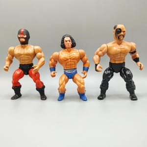 May include: Three vintage action figures with muscular physiques. The figures are in various poses and wear wrestling-themed attire. One figure wears a mask, another has long hair, and the third has face paint. The figures are made of plastic.