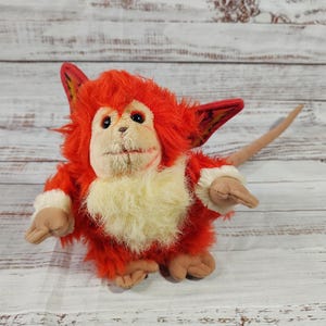May include: A plush toy creature with red fur, large ears, and a long tail. The toy has a cream-colored face and chest, with tan hands and feet. The creature's ears have a patterned design.