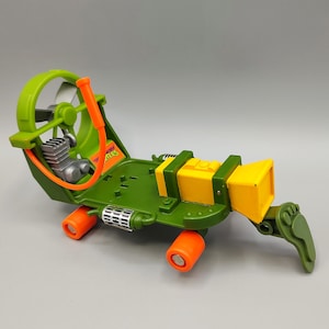May include: A green and yellow toy vehicle with orange accents. The toy has a fan, engine, and a foot-shaped element. The word "Turtles" is visible on the side. The vehicle has orange wheels.