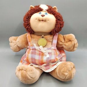 May include: A plush cat doll with brown fur, blue eyes, and a white patch on its head. It wears a brown collar with a round gold tag and a plaid jumper in shades of orange, pink, and white, featuring a small white bow and white lace trim.