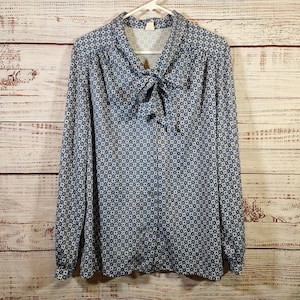 May include: A long-sleeved button-down blouse featuring a repeating geometric diamond pattern in shades of blue, white, and orange. The garment includes a pussy-bow collar detail at the neckline and is displayed on a white plastic hanger.