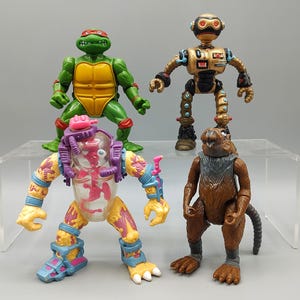 May include: Four vintage action figures: a green Teenage Mutant Ninja Turtle with a red mask, a gold robot, a mutant figure with a clear torso, and a brown anthropomorphic rodent. The figures are on a clear display shelf.