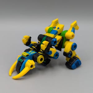 May include: A yellow, blue, black, and green toy robot with articulated joints and a claw-like appendage. The toy has a complex design with multiple components and a futuristic aesthetic. The robot is posed on a neutral gray surface.