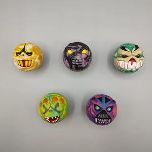 May include: Five colorful, round toys with monster faces. The toys feature various colors like yellow, purple, green, and blue, with detailed facial features including teeth, eyes, and expressions. The toys are arranged against a gray background.