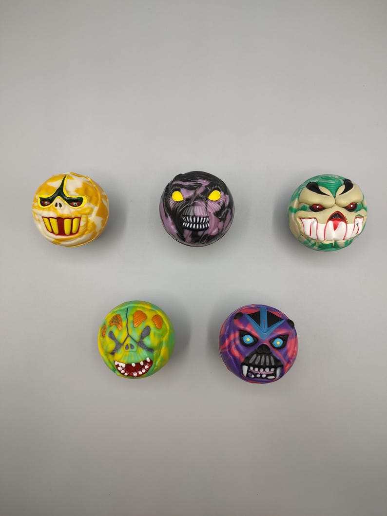 May include: Five colorful, round toys with monster faces. The toys feature various colors like yellow, purple, green, and blue, with detailed facial features including teeth, eyes, and expressions. The toys are arranged against a gray background.