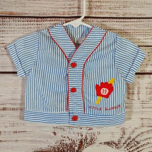 May include: Blue and white striped baby baseball shirt with red trim and buttons. Features a baseball glove and bat applique with the text "Little Slugger". Short-sleeved, button-down shirt hanging on a white hanger.