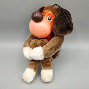 May include: A vintage plush dog toy with brown fur, white paws, and a large, orange and pink plastic nose. The dog has a red scarf and a black string for hanging. The toy is in a sitting position, with its arms wrapped around itself.